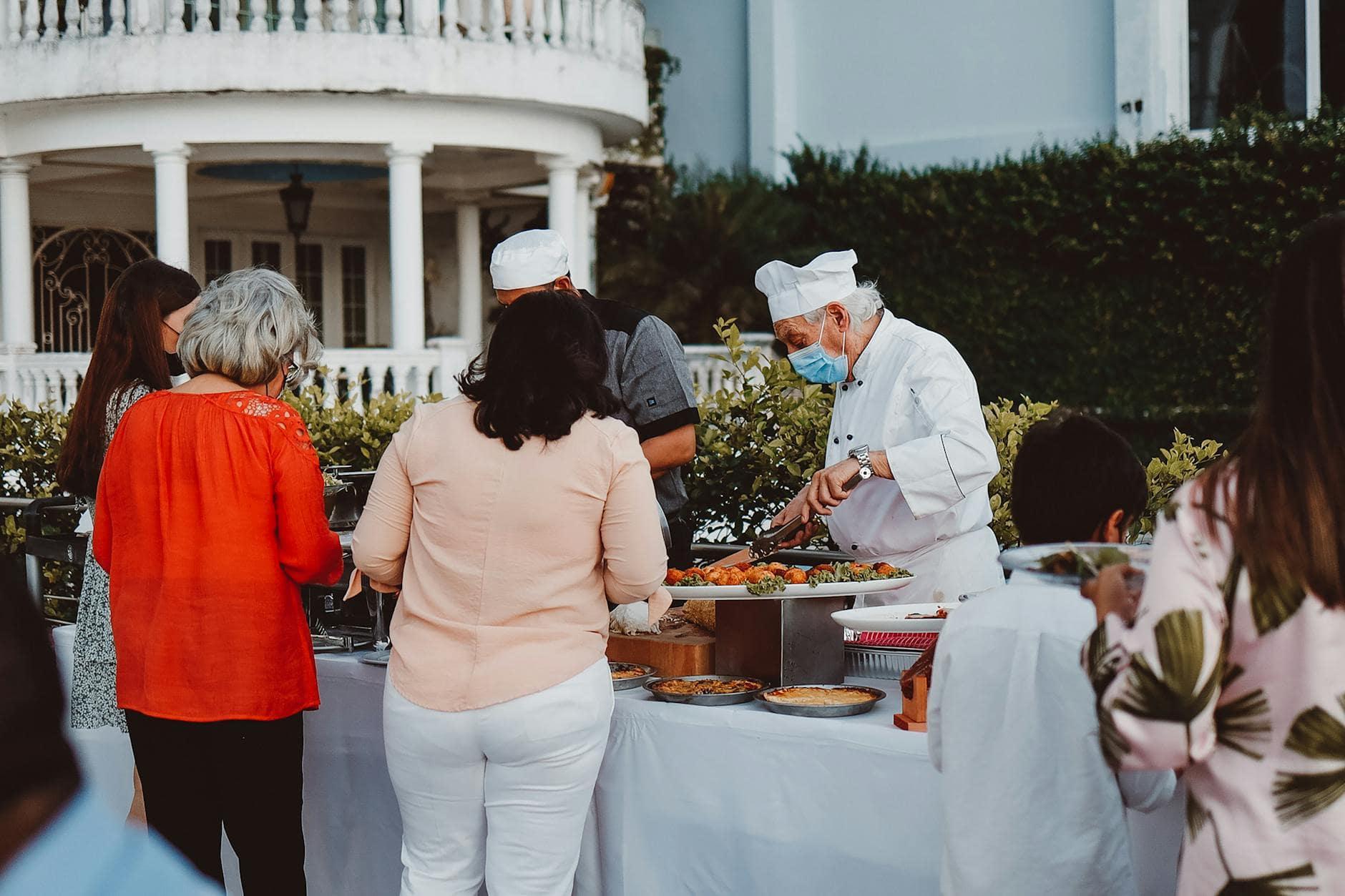 Inicio Chef with mask serves guests at outdoor catered event in elegant setting.
